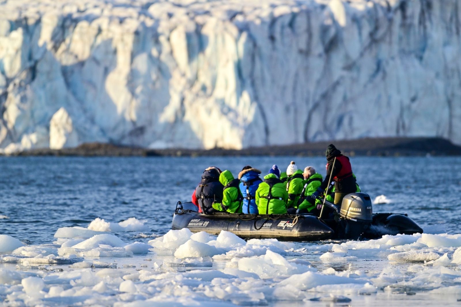 Travelers in bright expedition jackets riding in a Zodiac through icy water beneath a towering glacier front.